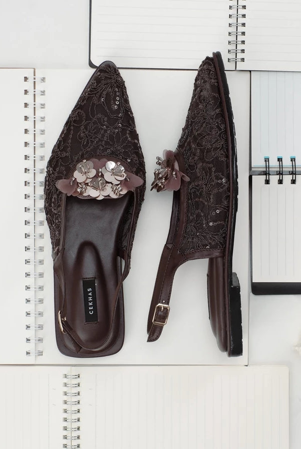 Pair of brown embellished sandals by Cekhas on a white surface with a notebook.