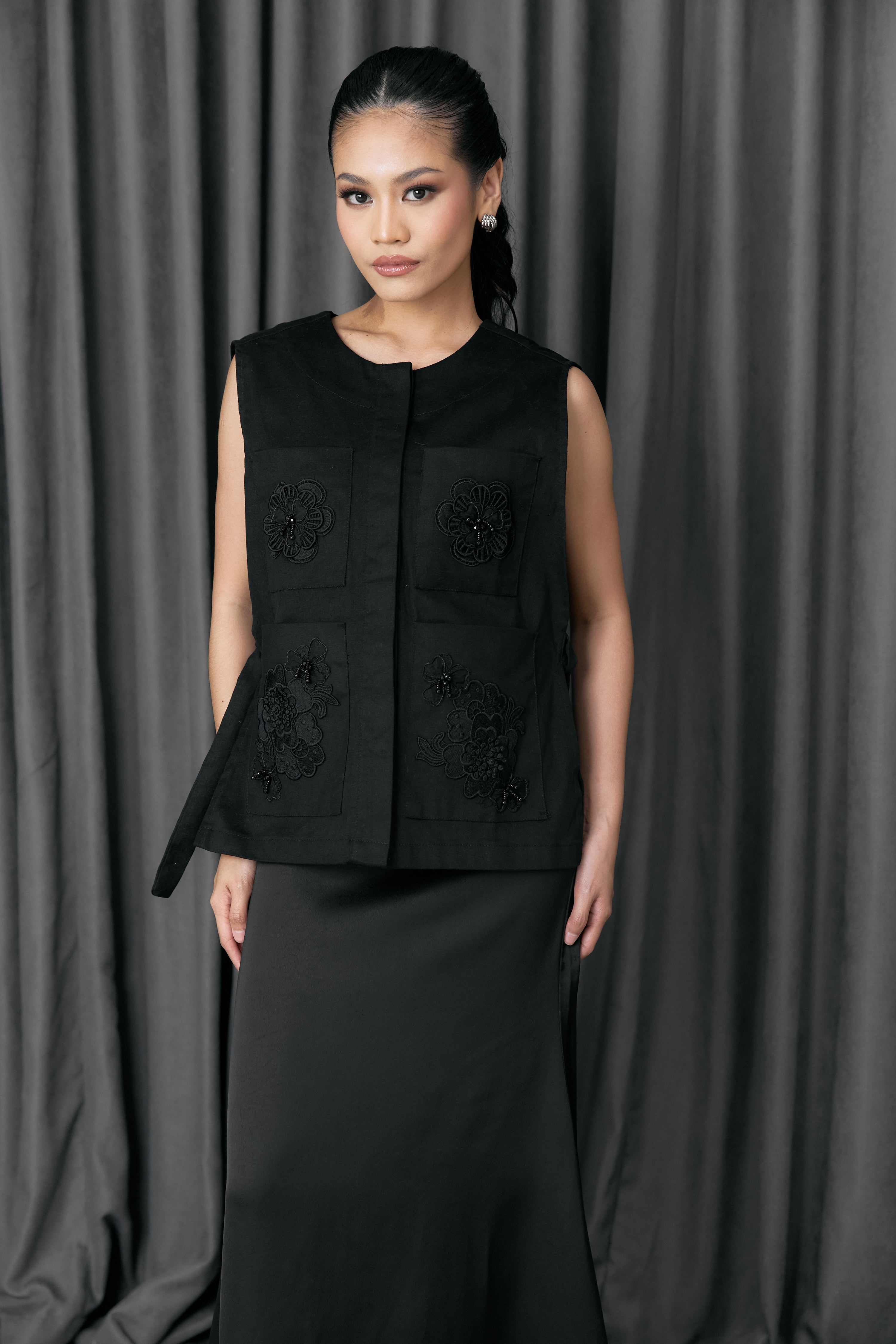 Woman wearing a black sleeveless vest with floral embroidery by Studio Tui against a gray curtain background