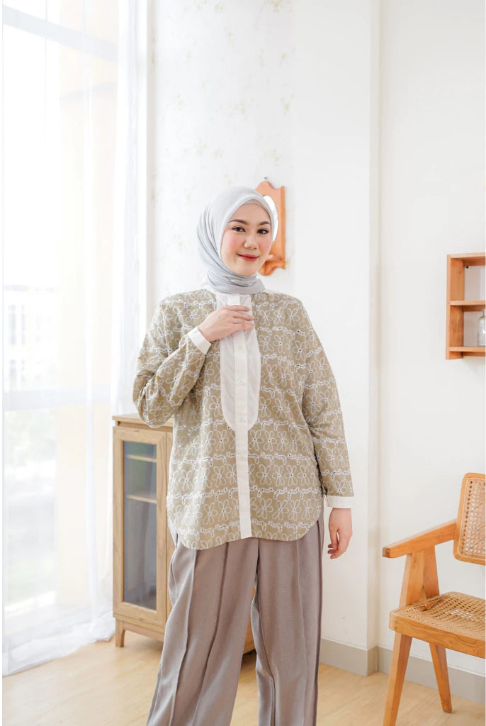 A woman wearing an olive green shirt by Abame with a long sleeves, paired with light brown trousers, standing in a room with a light decor.