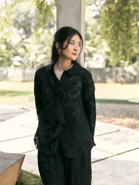 Woman in a black shirt with floral detail by Studio Tui standing outdoors with greenery in the background