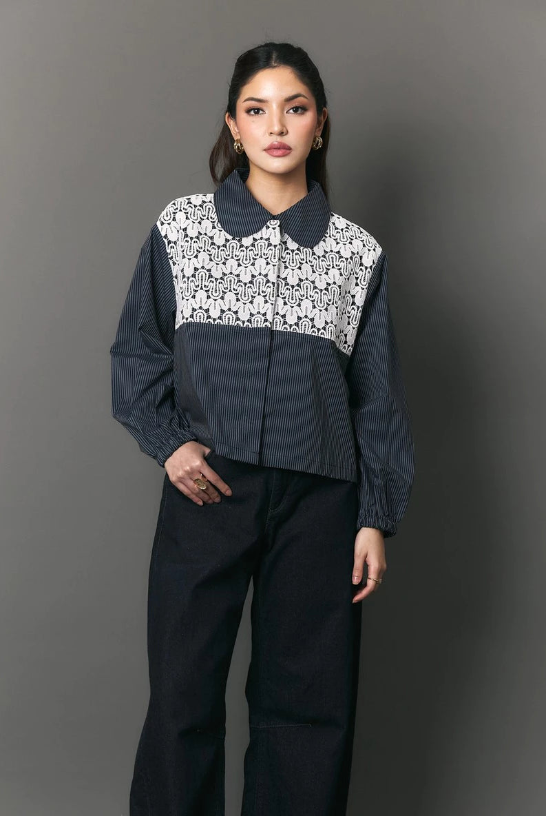 A model stands against a dark gray studio background wearing a cropped long-sleeve collared shirt by Studio Tui with a navy blue base and a white floral lace panel across the chest, paired with dark wide-leg pants.