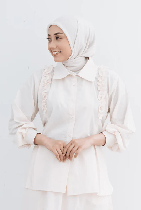 Woman wearing a white hijab and blouse with ruffled details from Atala Scarves on a light gray background