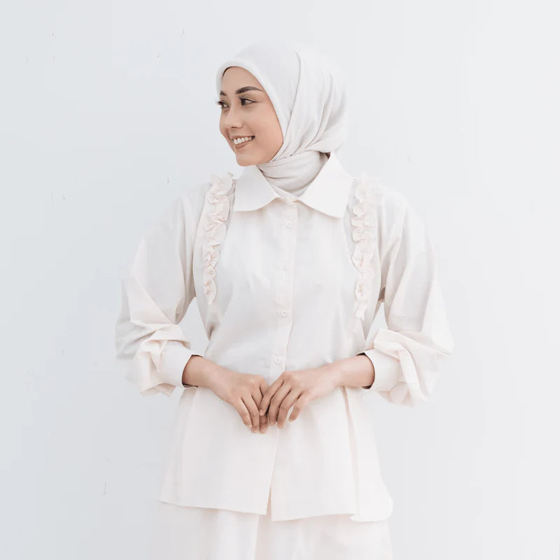 Woman wearing a white hijab and blouse with ruffled details from Atala Scarves on a light gray background