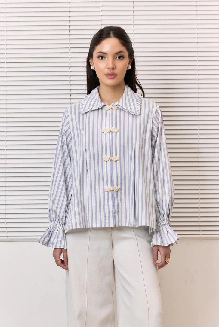 A woman stands in front of white window blinds wearing a light blue and white vertically striped shirt by Studio Tui with cream-colored decorative buttons and white trousers.