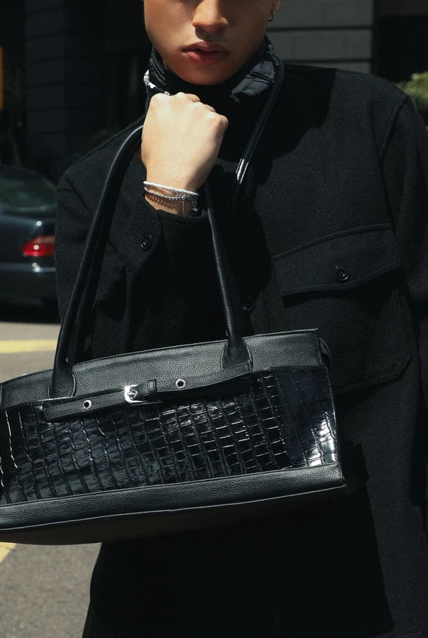 A person wearing a black coat carries a structured black handbag by Cekhas with a glossy crocodile-embossed front panel and double shoulder straps, photographed outdoors on a city street.