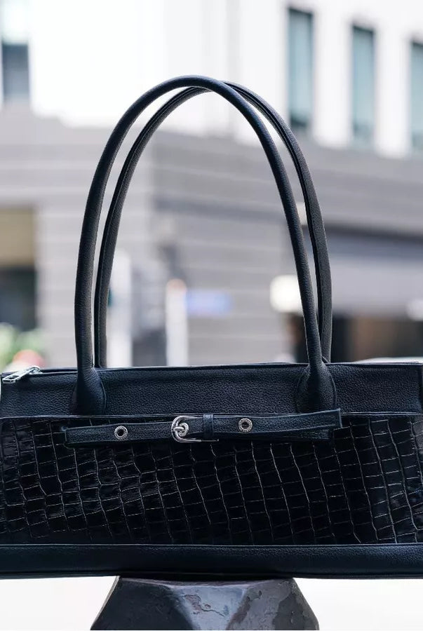 A black crocodile-embossed leather handbag by Cekhas with dual top handles and a front strap detail, displayed on a dark pedestal with a blurred urban street background.