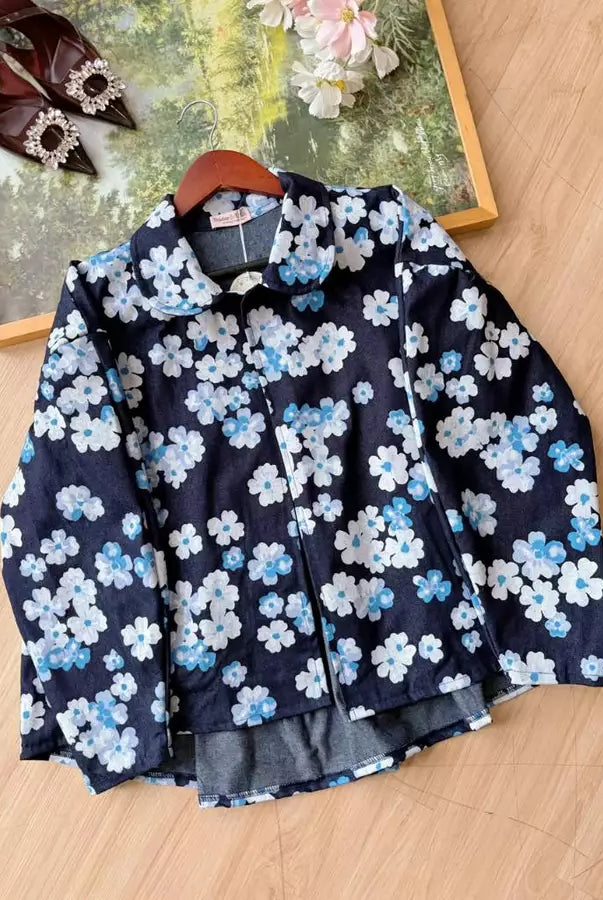 A denim jacket by Haidee Orlin with a navy blue base and a white and light blue floral print, displayed on a wooden hanger on a light wood floor next to a decorative frame.