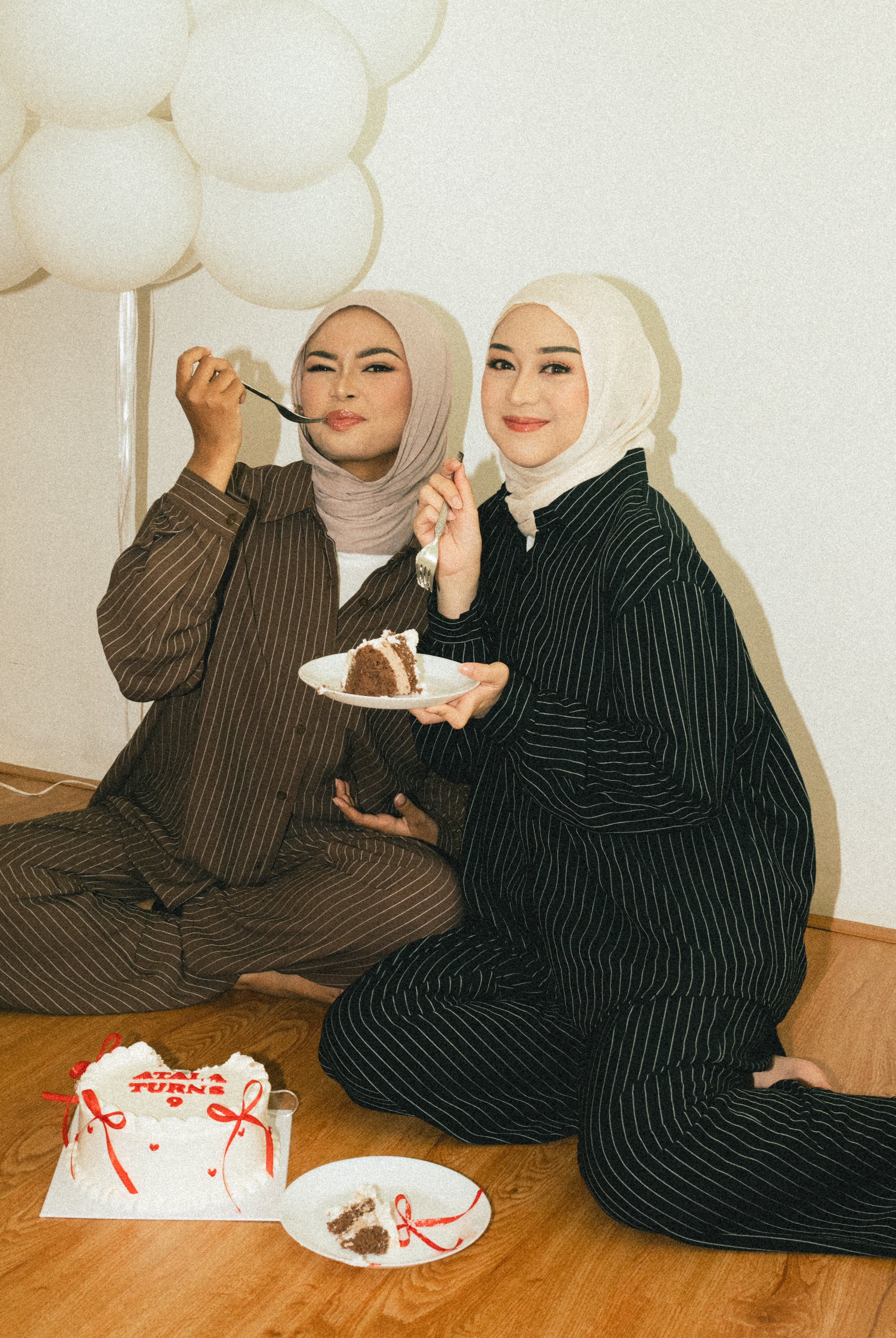 Two women in matching striped outfits from Atala Scarves sitting on the floor with balloons and a cake.