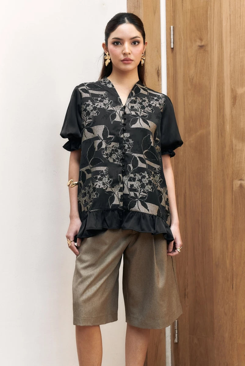 Woman wearing a black floral shortsleeves blouse by Studio Tui and beige shorts against a wooden door.