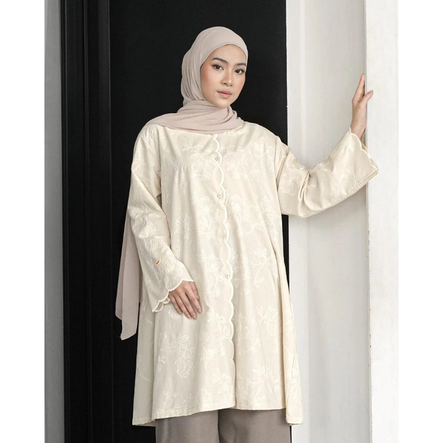 Woman wearing a cream-colored lace tunic by Mistyvori with a hijab against a black and white striped background.
Seorang wanita mengenakan tunik renda berwarna krem dari Mistyvori dengan hijab di latar belakang bergaris hitam-putih.