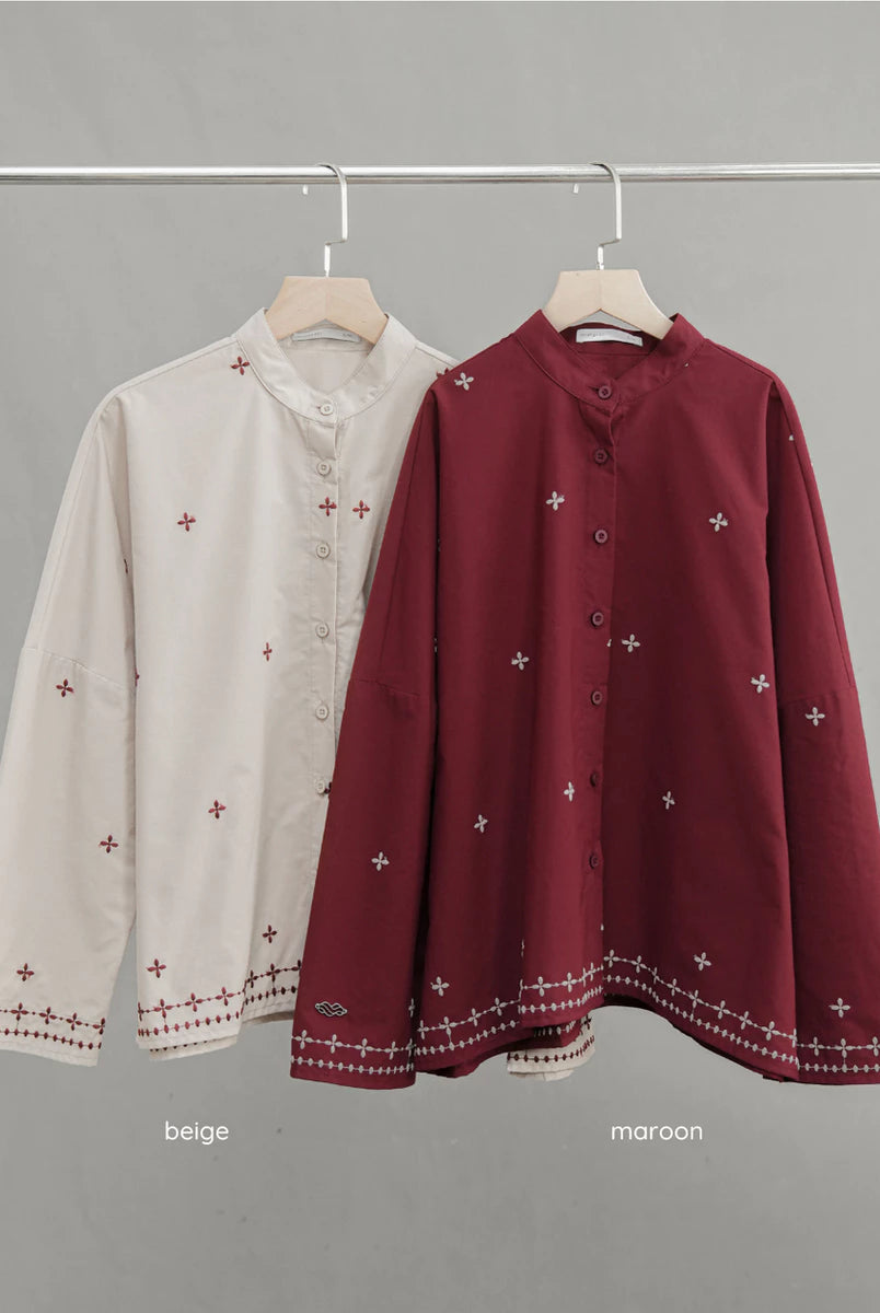 Two long-sleeve button-front blouses by Mistyvori hanging on a metal rack against a plain gray background. The left shirt is light beige with small red embroidered motifs and a red embroidered hem.