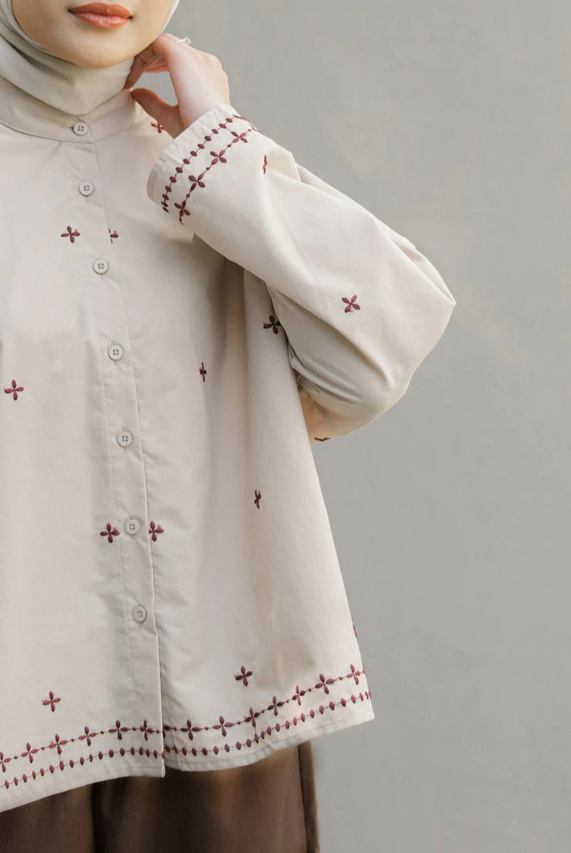 A close-up of a person wearing a cream-colored long-sleeve button-up blouse by Mistyvori with brown embroidered cross patterns and a matching hijab, set against a plain gray background.