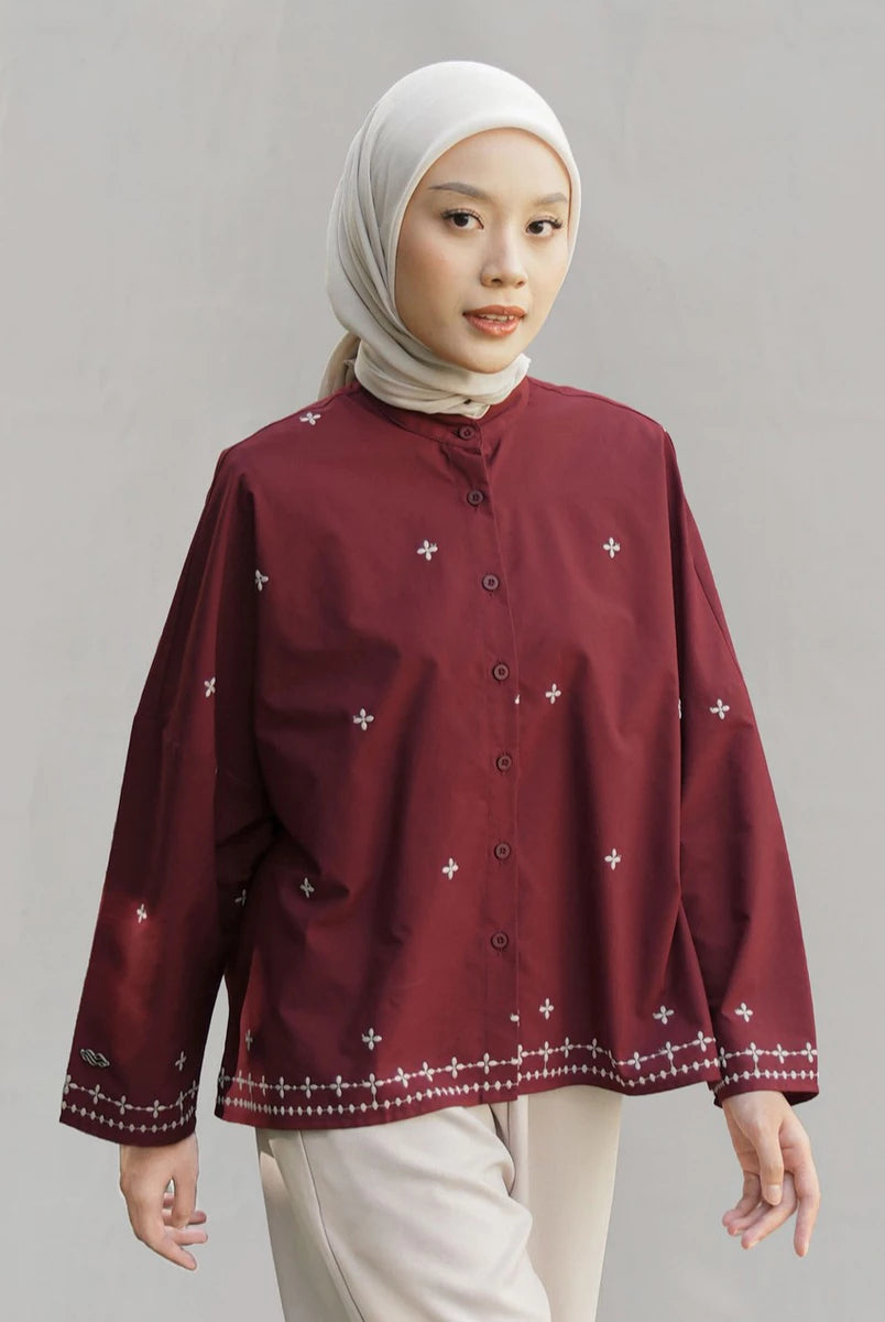 A woman wearing a maroon long-sleeve button-up blouse by Mistyvori with small white embroidered star motifs and a decorative white border along the hem and cuffs, paired with a light beige hijab and light beige trousers, set against a plain light gray studio background.