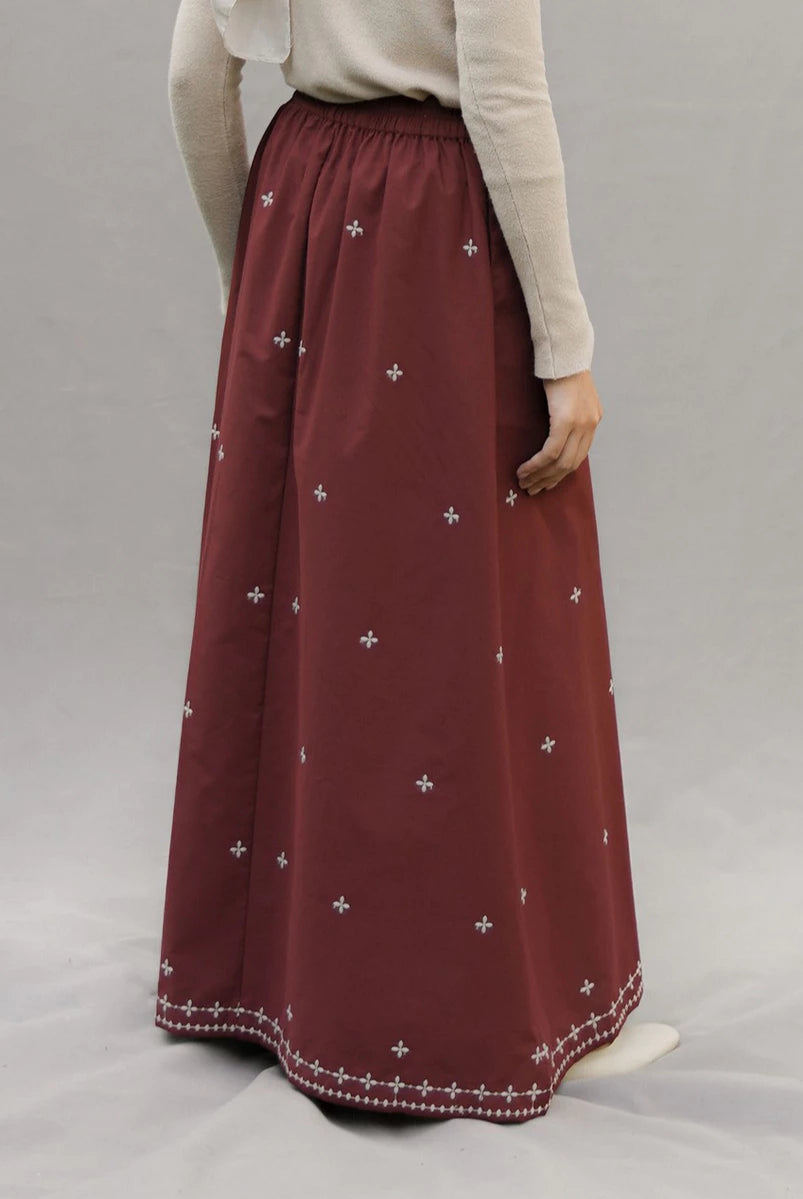 A person wearing a long pleated maroon skirt by Mistyvori with small white embroidered cross motifs and a decorative white border at the hem, paired with a light beige long-sleeve top against a neutral gray studio background.