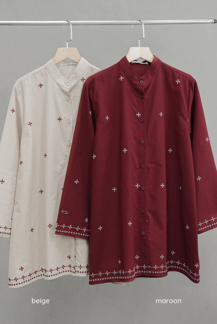 Two long-sleeve button-front tunic-style tops by Mistyvori hang on a metal rack against a plain gray wall. The front top is maroon with small white embroidered cross motifs and a decorative border near the cuffs and hem.