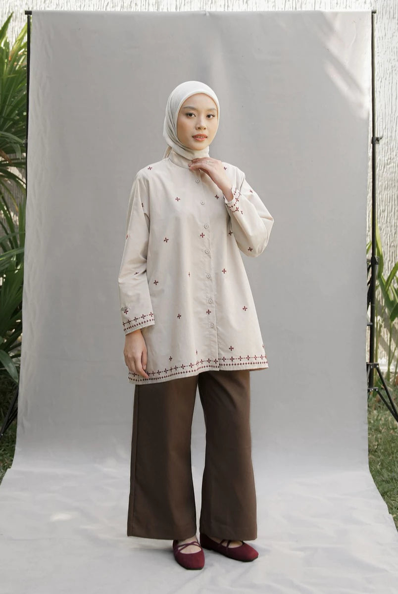 A woman wearing a cream-colored embroidered long-sleeve tunic by Mistyvori with brown wide-leg pants and a white hijab, standing in front of a white studio backdrop.