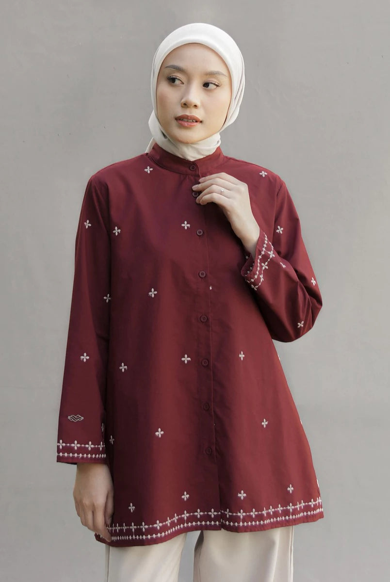 A woman wearing a maroon long-sleeve button-up tunic by Mistyvori with small white embroidered crosses and decorative borders, paired with a white hijab and light trousers, standing against a neutral gray studio background.