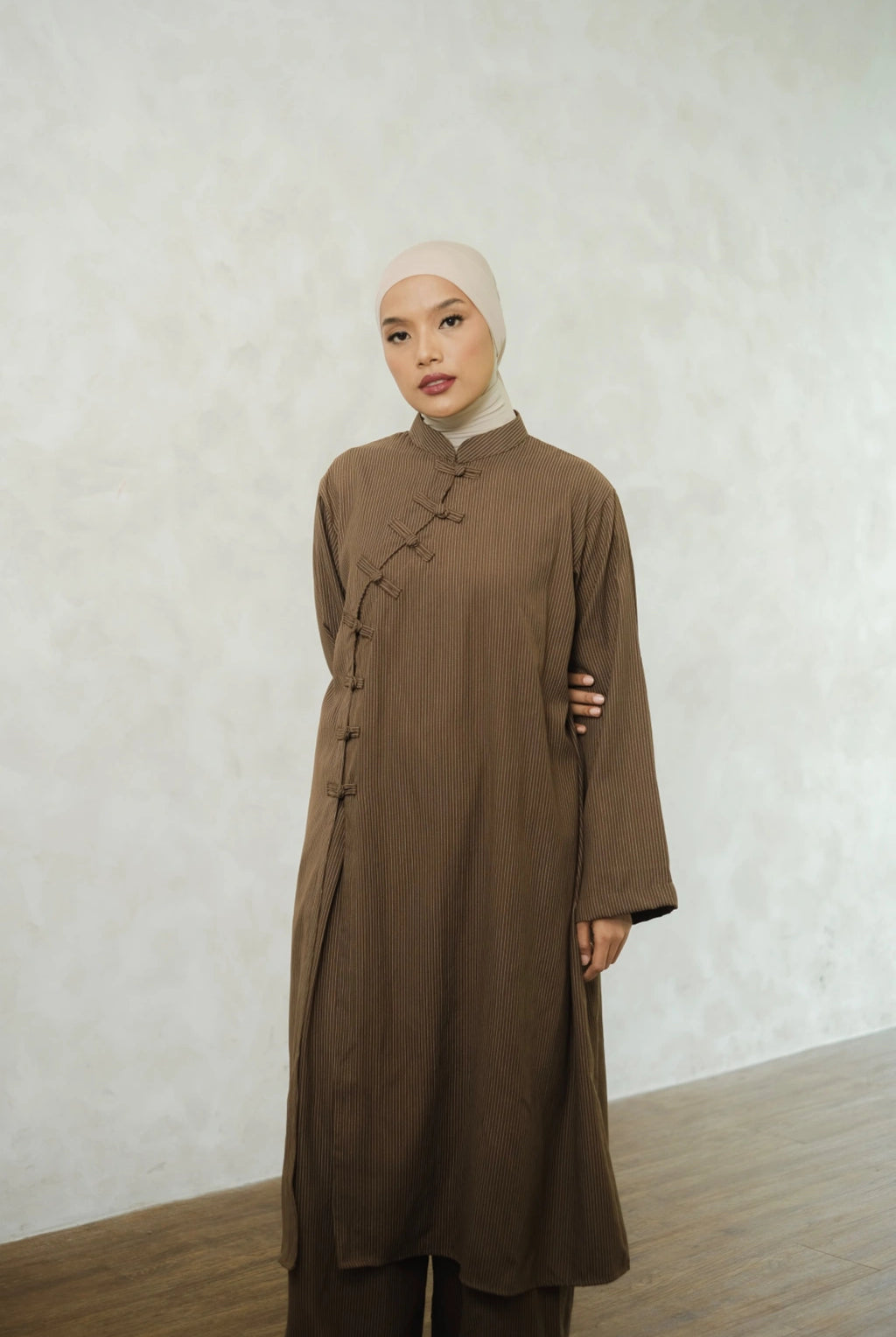 Woman wearing a brown long-sleeve tunic by Tufine and pants with a beige hijab against a white wall.
Seorang wanita mengenakan tunik lengan panjang berwarna cokelat dari Tufine dan celana panjang, serta hijab berwarna krem, dengan latar belakang dinding putih.