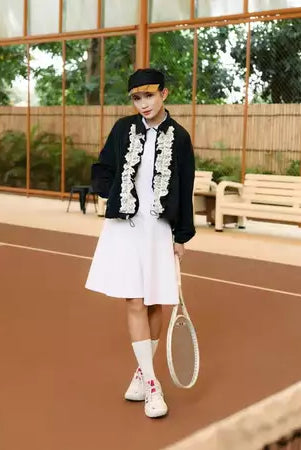 Person on a tennis court wearing a black jacket by Haidee Orlin with white floral embroidery and a white skirt.