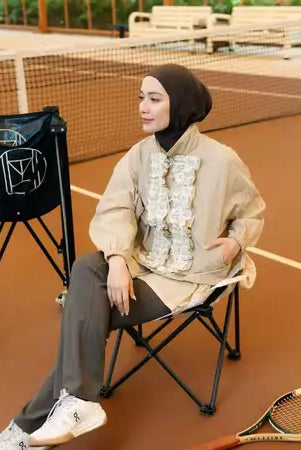 Person sitting on a chair on a tennis court wearing a beige jacket by Haidee Orlin with ruffled details.