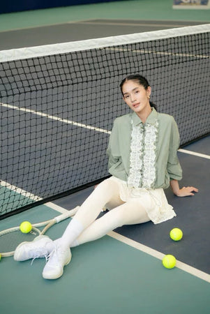 Woman sitting on a tennis court with tennis balls around her, wearing a sage green jacket by Haidee Orlin and white skirt.