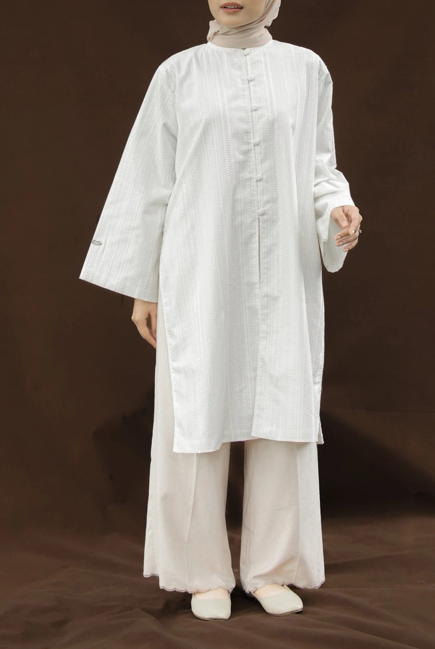 Woman wearing a white tunic by Mistyvori and a ivory pants on a brown background.
Seorang wanita mengenakan tunik putih dari Mistyvori dan celana panjang berwarna gading dengan latar belakang cokelat.