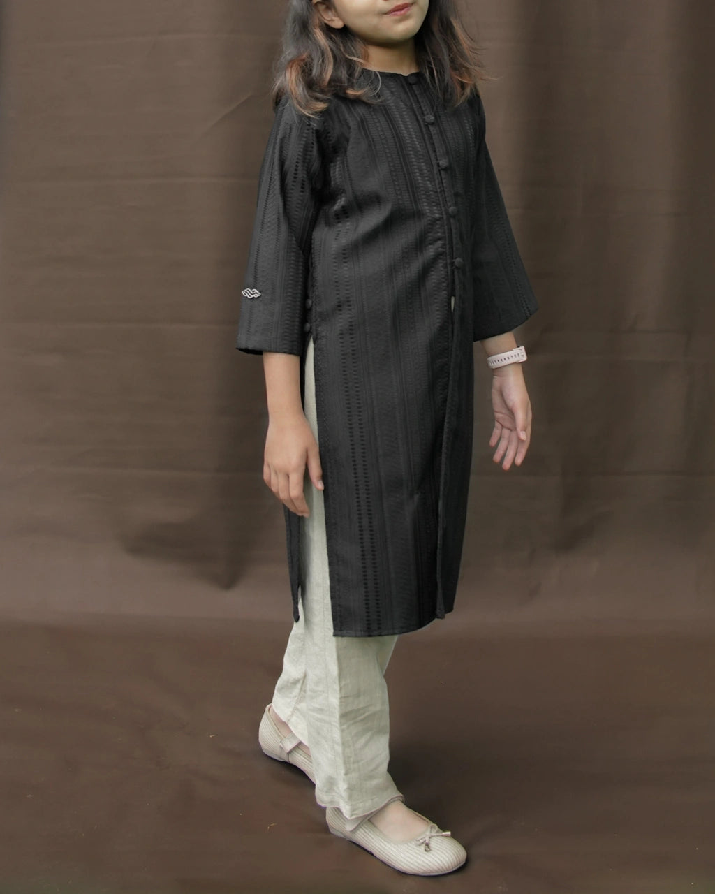 Child wearing a black tunic by Mistyvori with beige pants on a brown background.
Seorang anak mengenakan tunik hitam dari Mistyvori dengan celana krem di atas latar belakang cokelat.