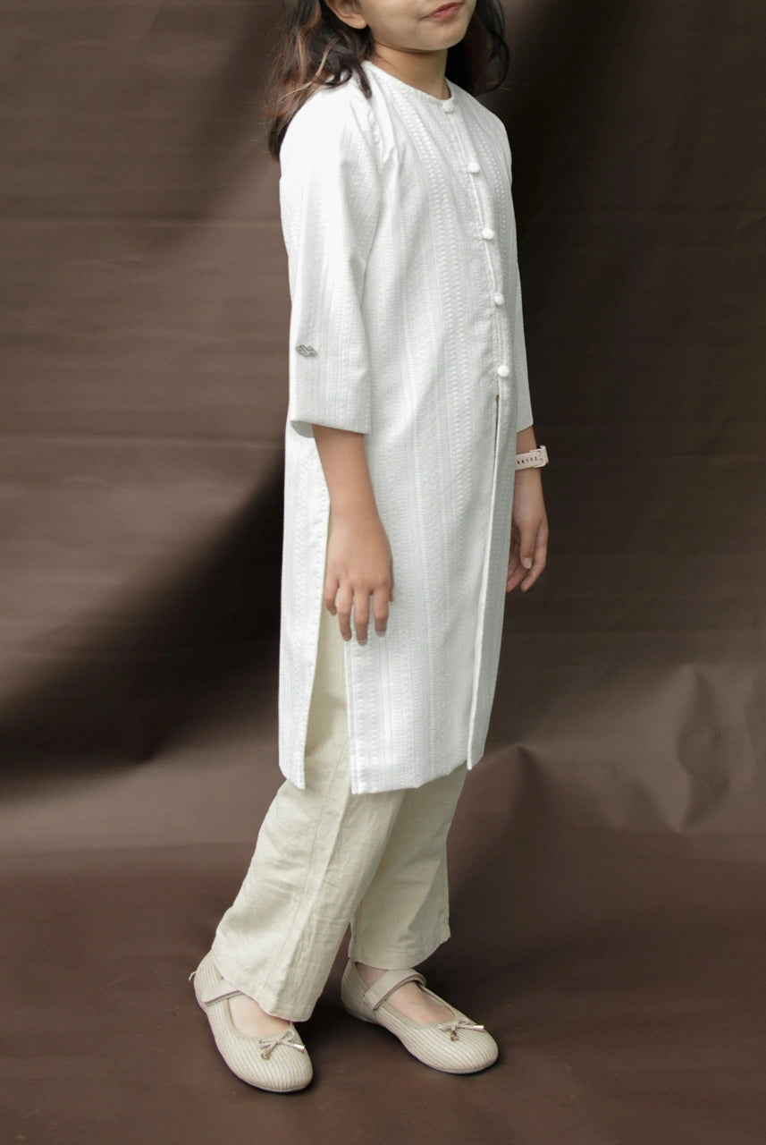 Child wearing a white tunic by Mistyvori with beige pants on a brown background.
Seorang anak mengenakan tunik putih dari Mistyvori dengan celana krem di atas latar belakang cokelat.
