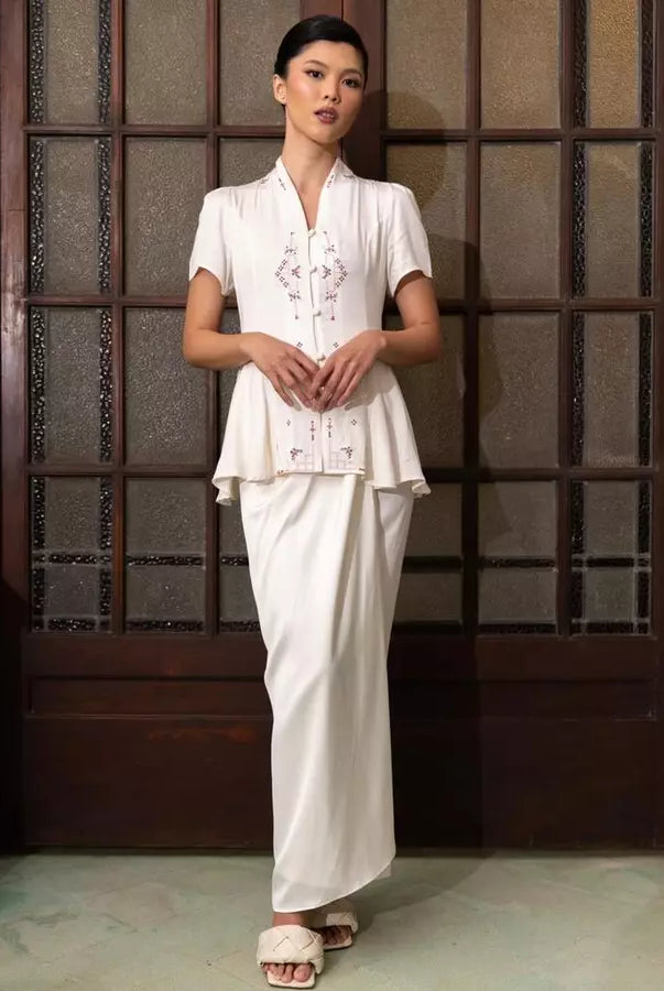 Woman wearing a traditional white kebaya by Kartinis Label standing against a wooden paneled wall.