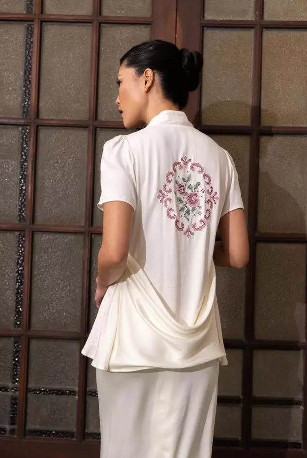 Woman wearing a white traditional kebaya by Kartinis Label with floral embroidery on the back, standing against a textured wall.