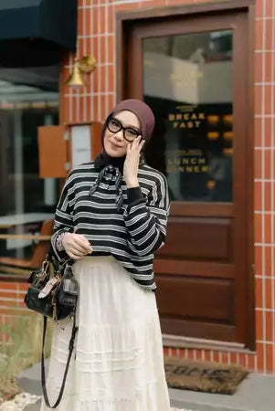 Woman in a striped knit sweaters by Haidee Orlin and hijab standing outside a building with a brick wall.