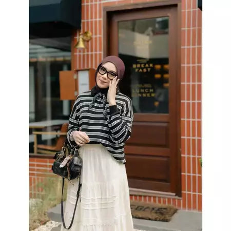 Woman in a striped knit sweaters by Haidee Orlin and hijab standing outside a building with a brick wall.
