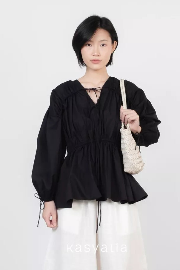 A model wearing a black long-sleeve blouse by Kasyalia with a V-neckline, tie details at the collar and cuffs, and a gathered peplum waist, paired with white drawstring pants and holding a cream woven shoulder bag.