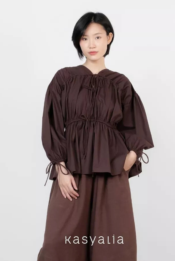 A model wearing a dark brown long-sleeve blouse with a gathered, tie-neck design and matching wide-leg trousers, set against a plain light gray background. The brand name 'kasyalia' is printed at the bottom.