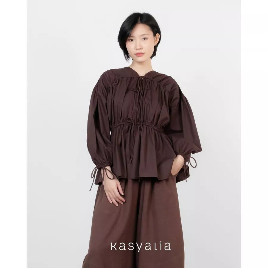 A model wearing a dark brown long-sleeve blouse with a gathered, tie-neck design and matching wide-leg trousers, set against a plain light gray background. The brand name 'kasyalia' is printed at the bottom.