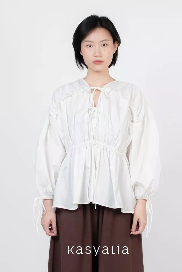 A model wearing a white long-sleeve blouse with a tie-front keyhole and gathered peplum waist, paired with dark brown wide-leg pants, against a plain light gray background. The brand name 'kasyalia' is printed at the bottom.