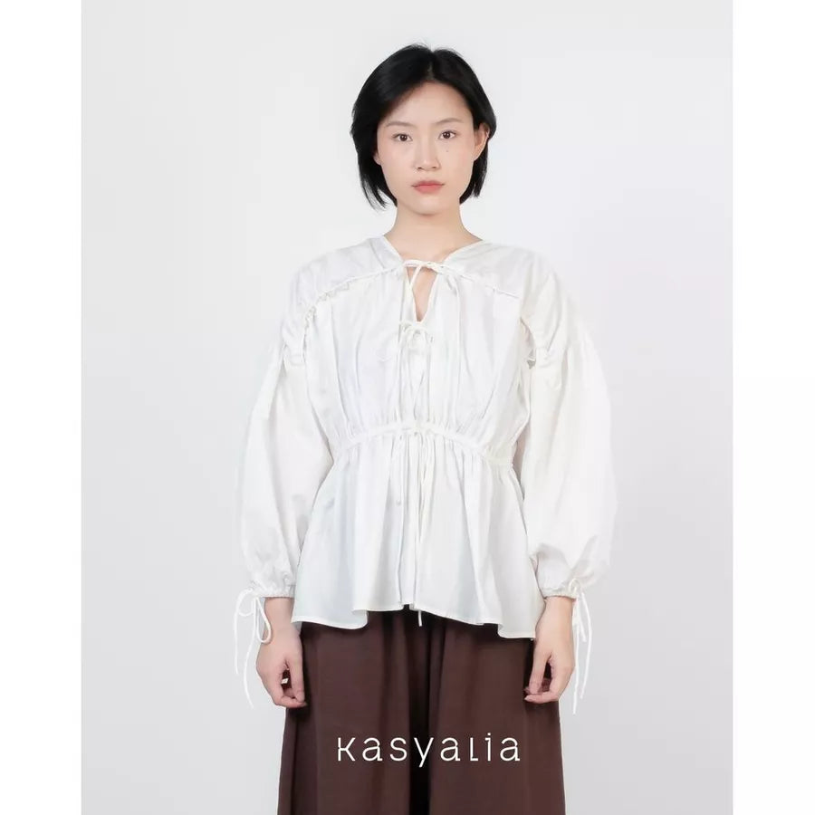 A model wearing a white long-sleeve blouse with a tie-front keyhole and gathered peplum waist, paired with dark brown wide-leg pants, against a plain light gray background. The brand name 'kasyalia' is printed at the bottom.