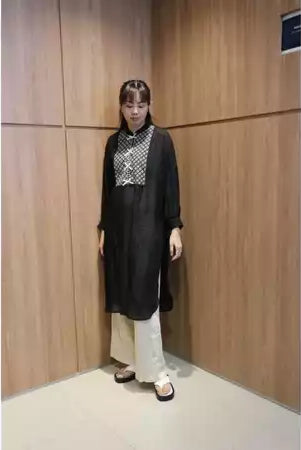 A person wearing a long black tunic with lace details, paired with light-colored pants and standing against a wooden backdrop.