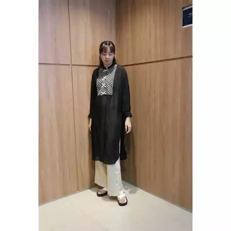 A person wearing a long black tunic with lace details, paired with light-colored pants and standing against a wooden backdrop.