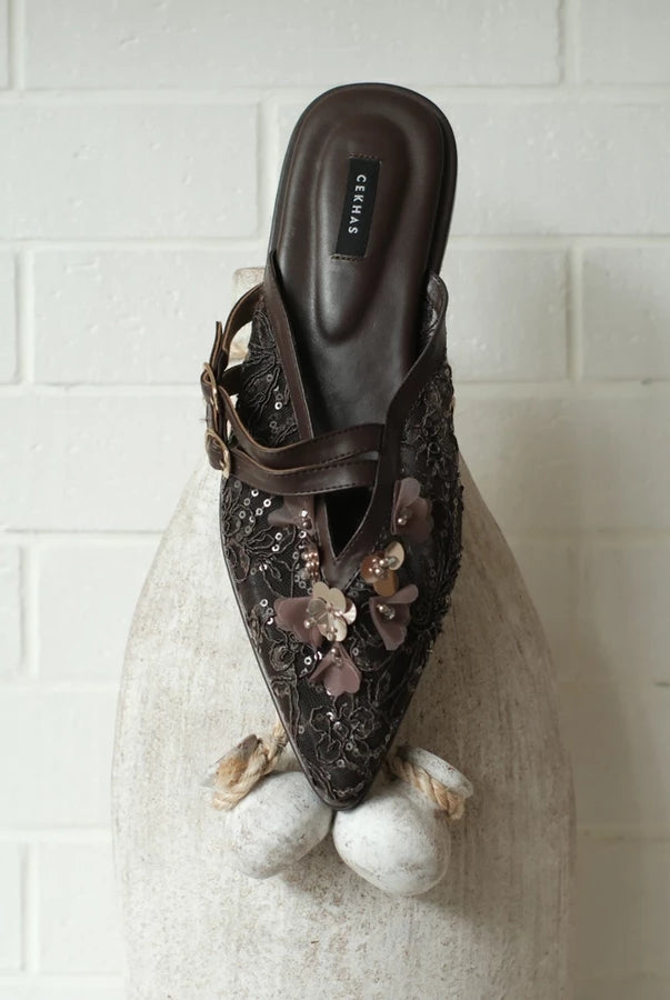 Brown mules sandal by Cekhas with floral embellishments on a textured surface with a white tiled background
