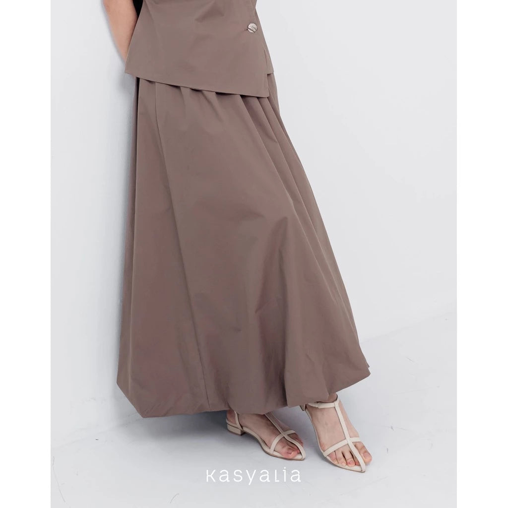 A woman wearing a brown Sienna skirt with soft pleats by Kasyalia, paired with a matching top. The skirt reaches down to the ankles and has a flowing silhouette.