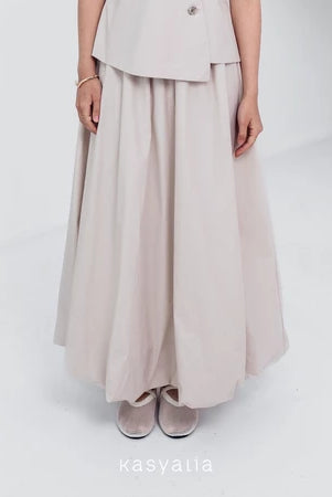 A woman wearing a cream Sienna skirt with soft pleats by Kasyalia, paired with a matching top. The skirt reaches down to the ankles and has a flowing silhouette.