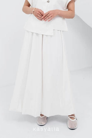 A woman wearing a white Sienna skirt with soft pleats by Kasyalia, paired with a matching top. The skirt reaches down to the ankles and has a flowing silhouette.
