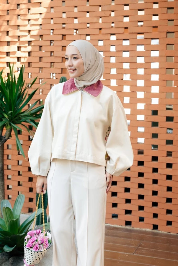 A woman wearing a cream-colored jacket by Haidee Orlin with a contrasting dusty pink collar, a beige hijab, and holding a small basket of pink flowers, standing in front of a terracotta lattice wall.