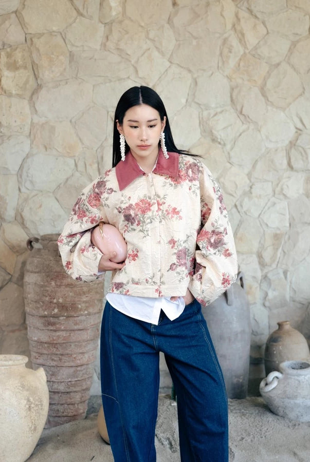 A woman stands in front of a rustic stone wall wearing a cream floral bomber jacket by Haidee Orlin with a dusty rose collar, dark blue wide-leg pants, and holding a pink clutch.