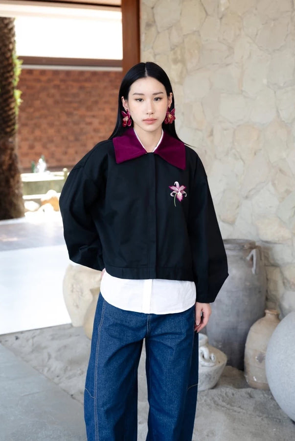 A woman stands in a stone-walled outdoor setting wearing a black cropped jacket by Haidee Orlin with a deep burgundy collar, a white shirt underneath, blue jeans, and large pink earrings.