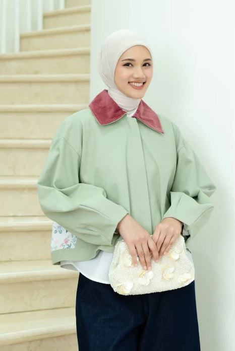 A woman wearing a sage green oversized long-sleeve jacket by Haidee Orlin with a contrasting dusty rose velvet collar, a white hijab, and dark navy pants, holding a white floral clutch bag in front of a white wall and beige staircase.