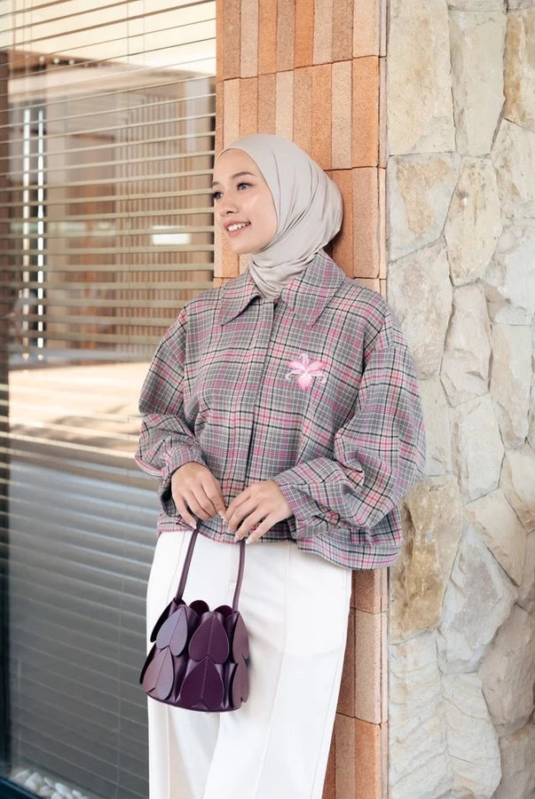 A woman wearing a light beige hijab and a pink and gray plaid long-sleeve jacket by Haidee Orlin with a pink bow accent, paired with white pants and holding a dark purple handbag, standing next to a stone wall.