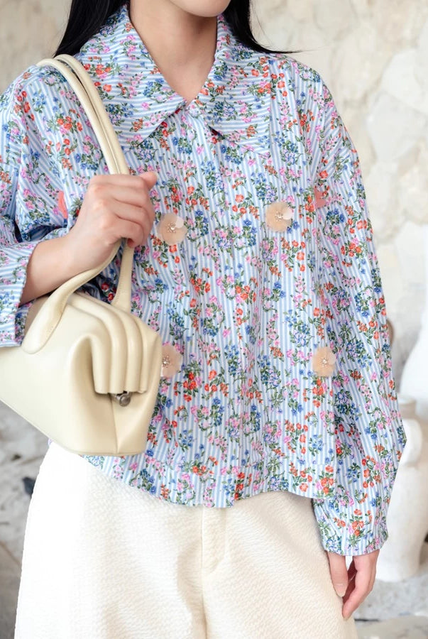 A person wearing a light blue and white floral long-sleeve jacket by Haidee Orlin with beige buttons decoration, paired with cream-colored pants and carrying a cream-colored shoulder bag.
