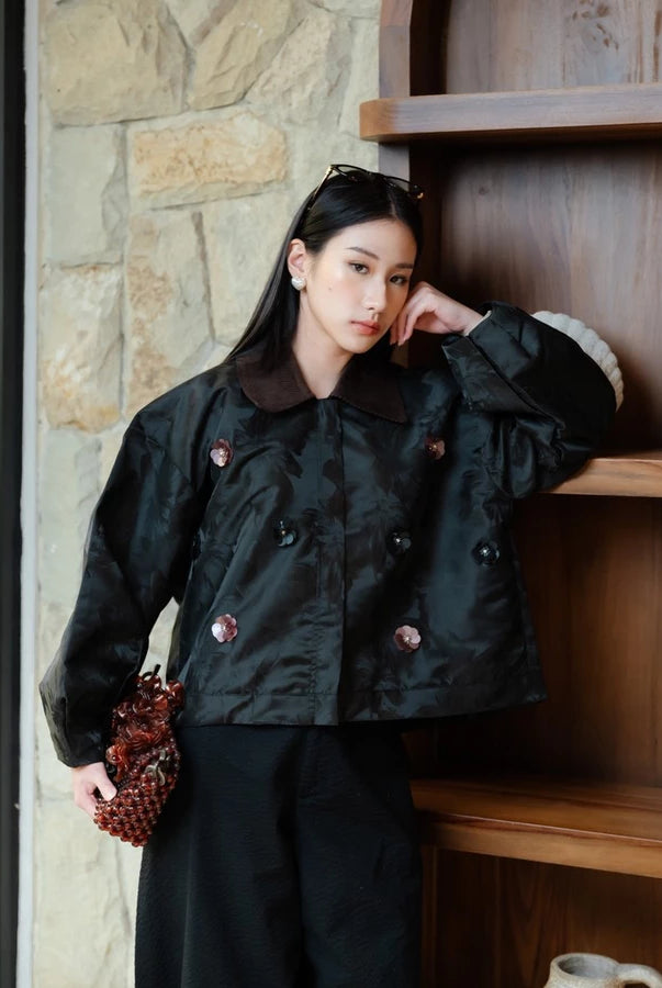 A woman models a black double-breasted jacket by Haidee Orlin with a dark brown collar and decorative buttons, paired with black trousers, standing next to a wooden shelf.
